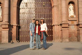 KC, Mirza, and Tiffiny is front of a large brown door with golden circles. The wall is made of brown stones. There are columns and archways. There are marble statues in the arches. KC (left) is wearing a blue shirt, jeans, glasses, and a brown baseball cap. She has a bag over her shoulder. Mirza is wearing a white shirt, red jacket, and jeans. She has shoulder length brown hair. Tiffiny is wearing a white shirt, a red jacket around her waist, jeans, and a white baseball cap.