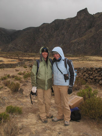 Tung- with a green jacket with the hood pulled up- and Catherine- wearing a blue jacket with the hood pulled up- are standing next to small rock walls. There are more walls in the background and a rocky hill in the background.