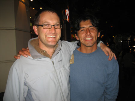 Steve and Abel are standing outside in the dark with their arms around each other. They are both smiling into the camera. There are various lights in the background. Steve is wearing a gray jacket with brown inside the collar, and black rimed glasses. Steve has very short brown hair. Abel is wearing a blue shirt. He has chin length black hair.