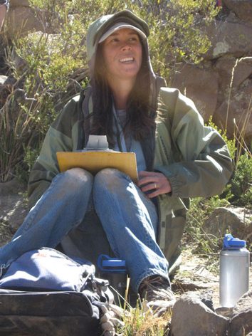 Tiffiny Tung is wearing jeans, a green jacket with the hood up, and a white baseball cap. She has long brown hair. She is in the field- sitting on the ground by rocks, shrubs, and water bottles. She is holding a clipboard and smiling to the right of the camera.